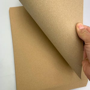 Peut inclure: Une pile de feuilles de papier kraft brun. Une feuille est soulevée pour révéler la texture et la couleur du papier. Le papier a un aspect naturel, non blanchi, adapté à divers travaux manuels et emballages.