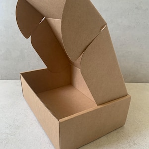 May include: An open, unfolded brown cardboard box. The box is rectangular with a lid that folds over. The box is made of corrugated cardboard and is sitting on a light gray surface.