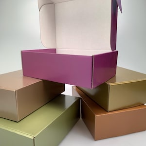 Könnte beinhalten: Eine Auswahl an glänzenden Pappschachteln in verschiedenen Farben. Die Schachteln sind in Lila, Gold, Bronze und Grün gehalten. Eine Schachtel ist geöffnet und zeigt einen weißen Innenraum. Die Schachteln sind aufeinander gestapelt.