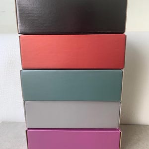 Può includere: Una pila verticale di cinque scatole di cartone rettangolari. La scatola in alto è nera, seguita da una scatola rossa, una scatola verde acqua spento, una scatola grigio chiaro e una scatola magenta in basso. Ogni scatola ha una finitura liscia.