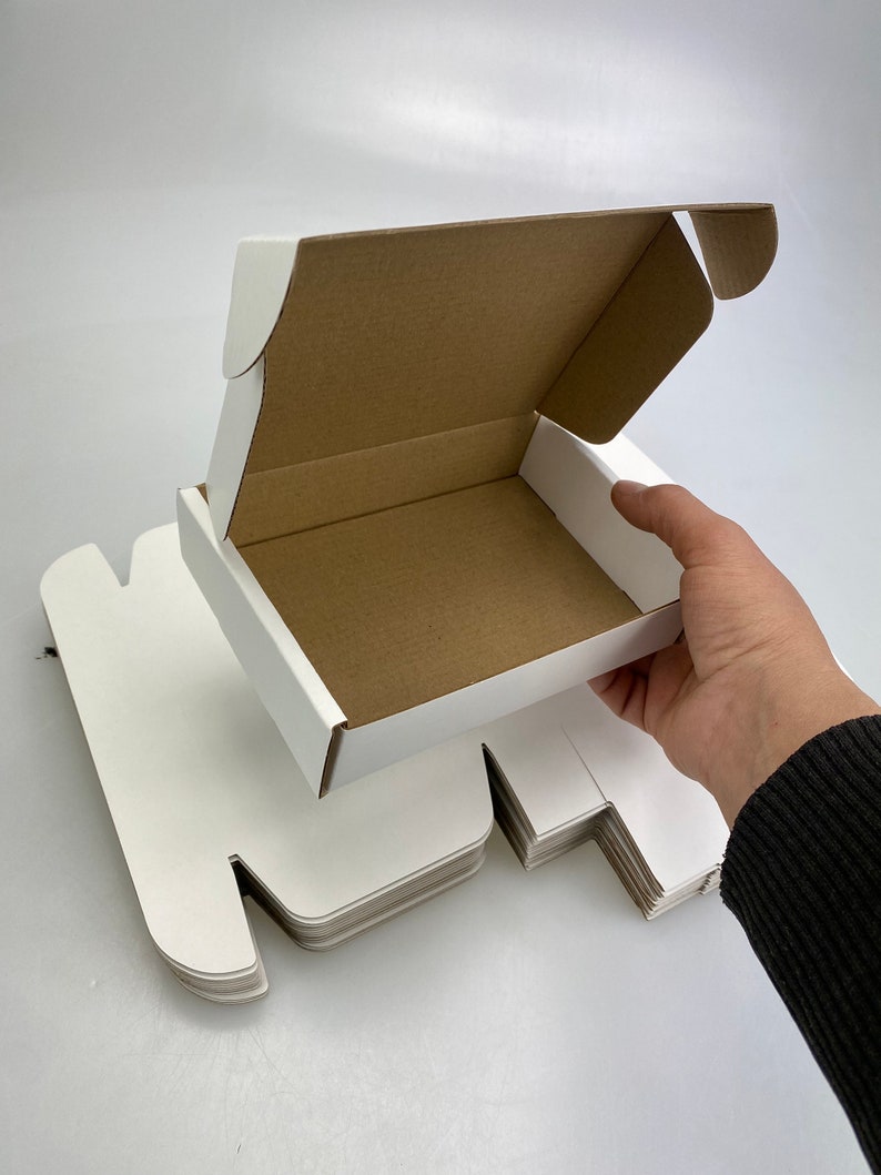 May include: A stack of white cardboard boxes with one box open, revealing a brown interior. The box is held open by a hand.