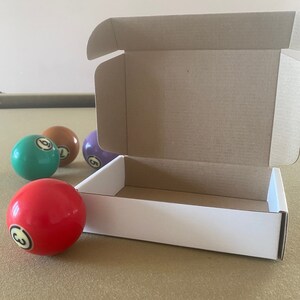 May include: A white cardboard box with a brown cardboard lid is open and sitting on a green felt surface. There are four billiard balls next to the box, one red, one green, one purple, and one orange.