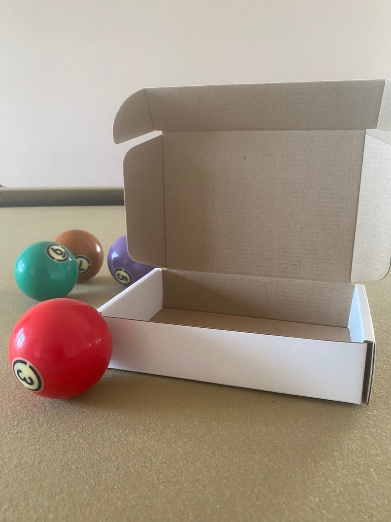 May include: A white cardboard box with a brown cardboard lid is open and sitting on a green felt surface. There are four billiard balls next to the box, one red, one green, one purple, and one orange.
