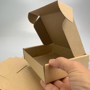 Könnte beinhalten: Eine kleine, offene, braune Pappschachtel mit einem gefalteten Deckel, die von einer Hand gehalten wird. Mehrere flache, ungefaltete Pappstücke sind in der unteren linken Ecke gestapelt. Die Schachtel ist leer, und der Hintergrund ist einfarbig weiß.
