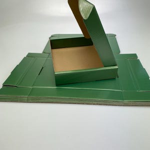 Op de afbeelding: Groene golfkartonnen dozen, waarvan één open, gestapeld op een wit oppervlak. De open doos onthult een bruin interieur. De dozen zijn gevouwen en klaar voor gebruik. De dozen hebben een effen groene kleur.