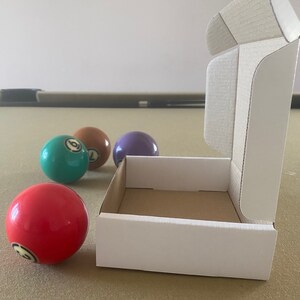 May include: A white cardboard box with a brown interior is open and sitting on a brown felt surface. Four billiard balls are visible, one red, one green, one purple, and one brown.