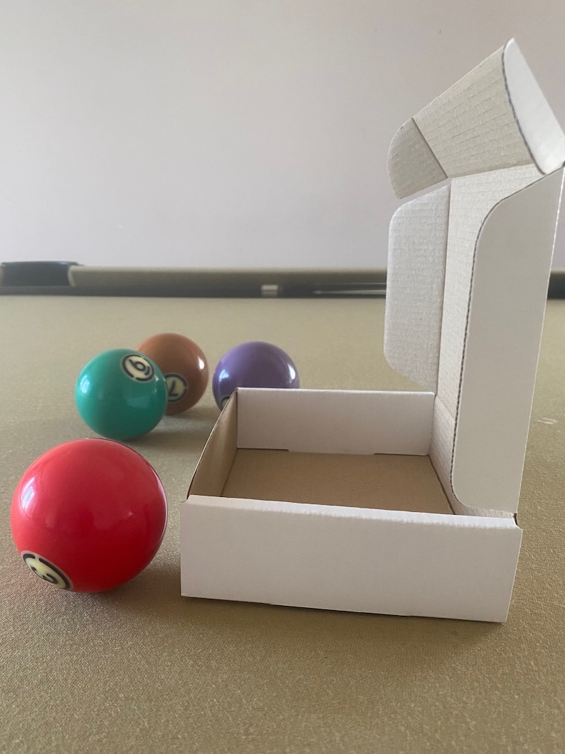 May include: A white cardboard box with a brown interior is open and sitting on a brown felt surface. Four billiard balls are visible, one red, one green, one purple, and one brown.