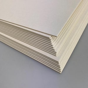 Pode incluir: Uma pilha de folhas de papel retangulares branco-sujo. As bordas revelam uma estrutura em camadas com um núcleo castanho claro. O papel está empilhado sobre uma superfície cinza, com as folhas superiores em foco.