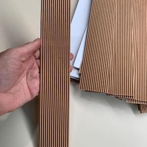 Könnte beinhalten: Ein hellbraunes, vertikales Wellpappelement, das von einer Hand gehalten wird, mit einem Stapel ähnlicher Elemente im Hintergrund. Die Elemente weisen vertikale Rillen auf. Der Hintergrund ist neutral, cremefarben. Geeignet für Verpackung oder Bastelarbeiten.