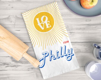 May include: White kitchen towel with a yellow sun and the word "LOVE" in white letters. The sun has rays coming out of it. Below the sun, it says "FROM Philly" in blue letters.