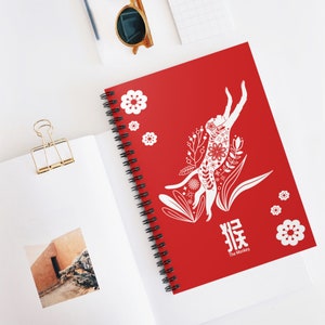 May include: A red spiral notebook with a white illustration of a monkey jumping in a field of flowers. The text "猴 The Monkey" is printed below the illustration.