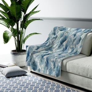 May include: A soft blue and white throw blanket with a watercolor wave pattern. The blanket is draped over a light gray couch with a green plant in a pot in the foreground.