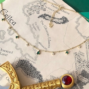 Puede incluir: Un collar de oro con una cadena delicada y pequeñas piedras preciosas verdes y transparentes. El collar está sobre un mapa con las palabras "Crilea" y "The Western Wastes" visibles.
