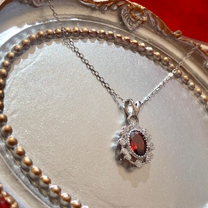 May include: Silver necklace with a pendant featuring a large oval, deep red gemstone surrounded by small, clear gemstones. The pendant hangs from a delicate silver chain, displayed on a silver tray with a gold beaded edge.