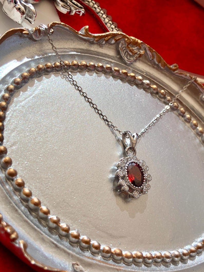 May include: Silver necklace with a pendant featuring a large oval, deep red gemstone surrounded by small, clear gemstones. The pendant hangs from a delicate silver chain, displayed on a silver tray with a gold beaded edge.