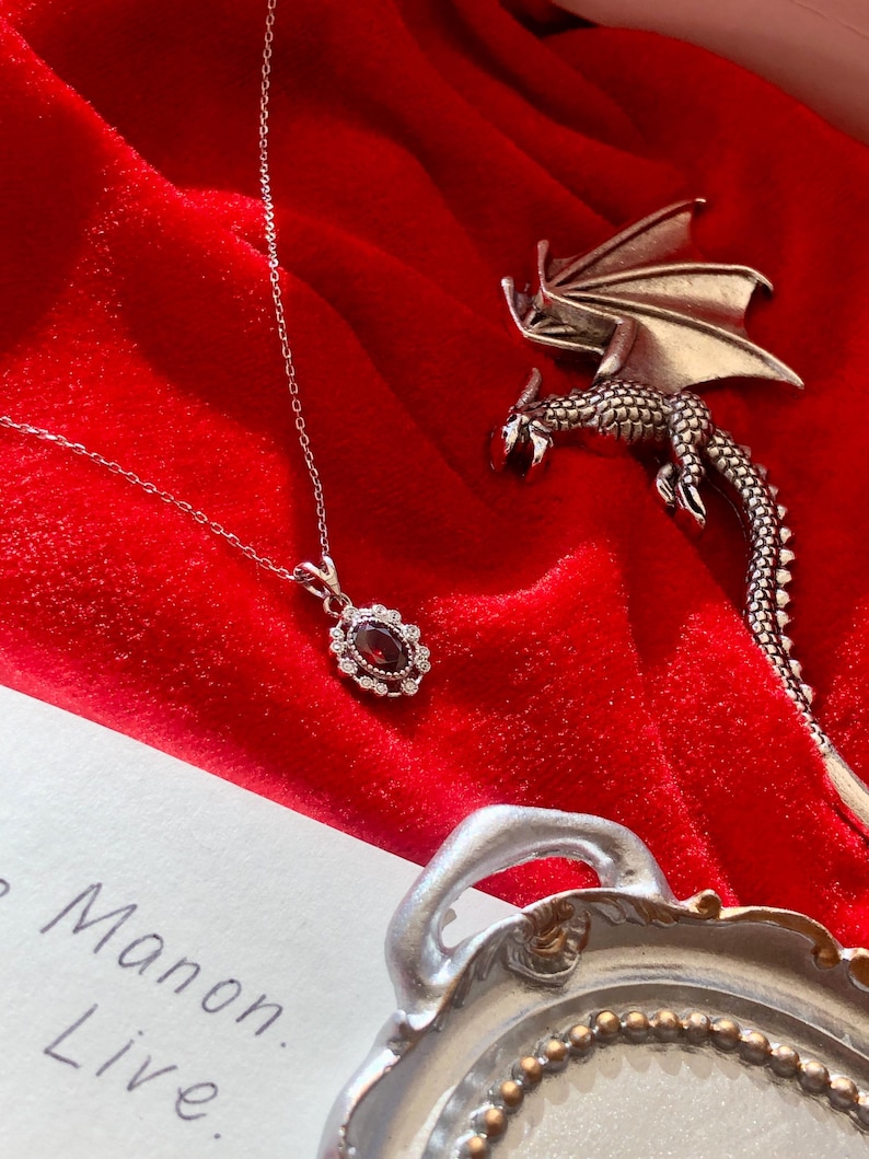 May include: A silver necklace with a dark red oval pendant surrounded by small white stones. A silver dragon brooch and a decorative silver tray are also on a red velvet surface. The text "Manon. Live." is visible.