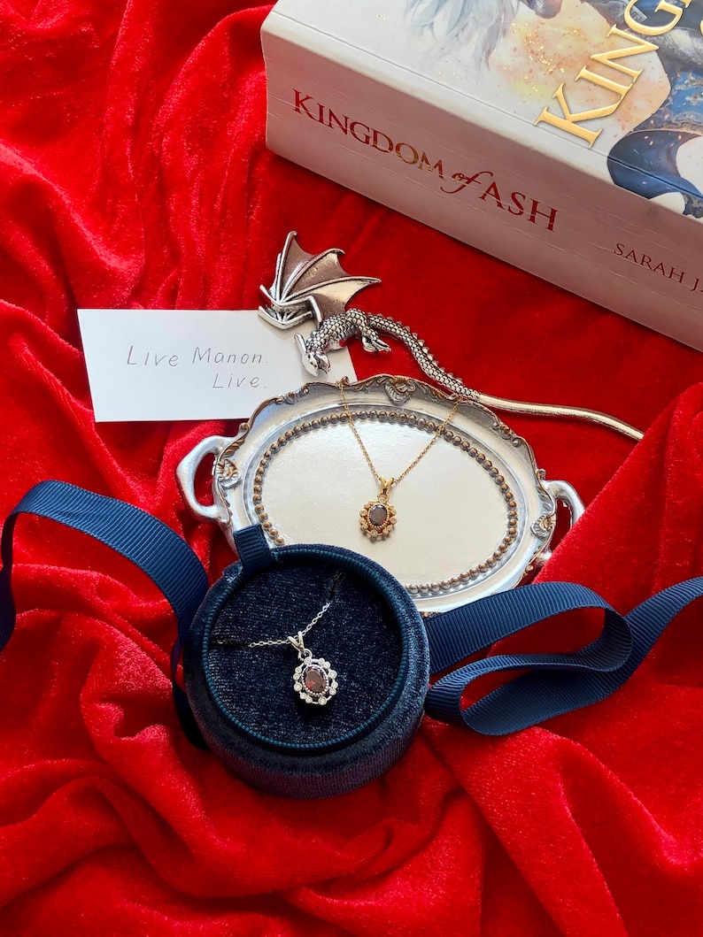 May include: A silver necklace with a dark gemstone pendant in a velvet box, displayed on a red velvet surface. A second necklace rests on a silver tray. A book titled "Kingdom of Ash" is visible in the background. The text "Live Manon Live" is on a card.