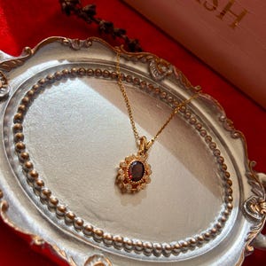 May include: A gold-colored necklace with a pendant featuring a dark red oval stone surrounded by small, clear stones. The necklace is displayed on a decorative silver tray with a beaded edge, set against a red background. The word "FASH" is visible in the background.