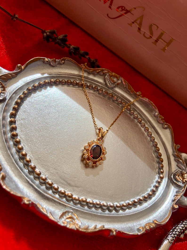 May include: A gold-colored necklace with a pendant featuring a dark red oval stone surrounded by small, clear stones. The necklace is displayed on a decorative silver tray with a beaded edge, set against a red background. The word "FASH" is visible in the background.