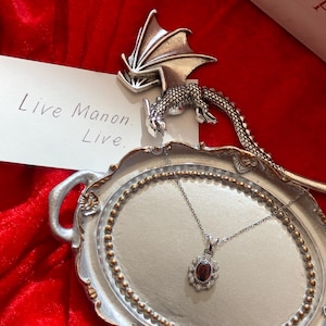 May include: A silver-toned necklace with a dark red oval gemstone pendant, surrounded by smaller clear stones, rests on a decorative silver tray. A silver dragon figurine and a card with the text "Live Manon. Live." are also present, set against a red velvet backdrop.