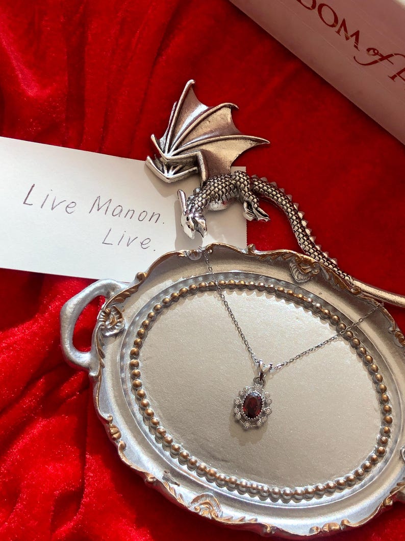 May include: A silver-toned necklace with a dark red oval gemstone pendant, surrounded by smaller clear stones, rests on a decorative silver tray. A silver dragon figurine and a card with the text "Live Manon. Live." are also present, set against a red velvet backdrop.