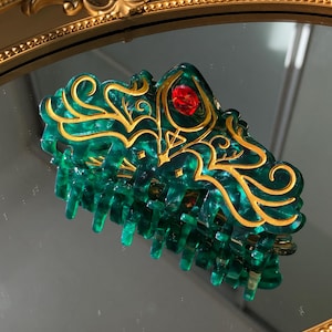 Könnte beinhalten: Grüne und goldene Haarspange mit dekorativem Design. Der Clip hat einen roten Edelstein und goldene Akzente. Die Haarspange besteht aus einem durchscheinenden grünen Material und ist dafür konzipiert, das Haar an Ort und Stelle zu halten.