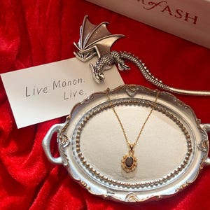 May include: A gold necklace with a dark oval pendant, surrounded by small white stones, rests on a silver tray. A silver dragon-shaped bookmark and a white card with the text "Live Manon. Live." are also present. The background is a red velvet surface.