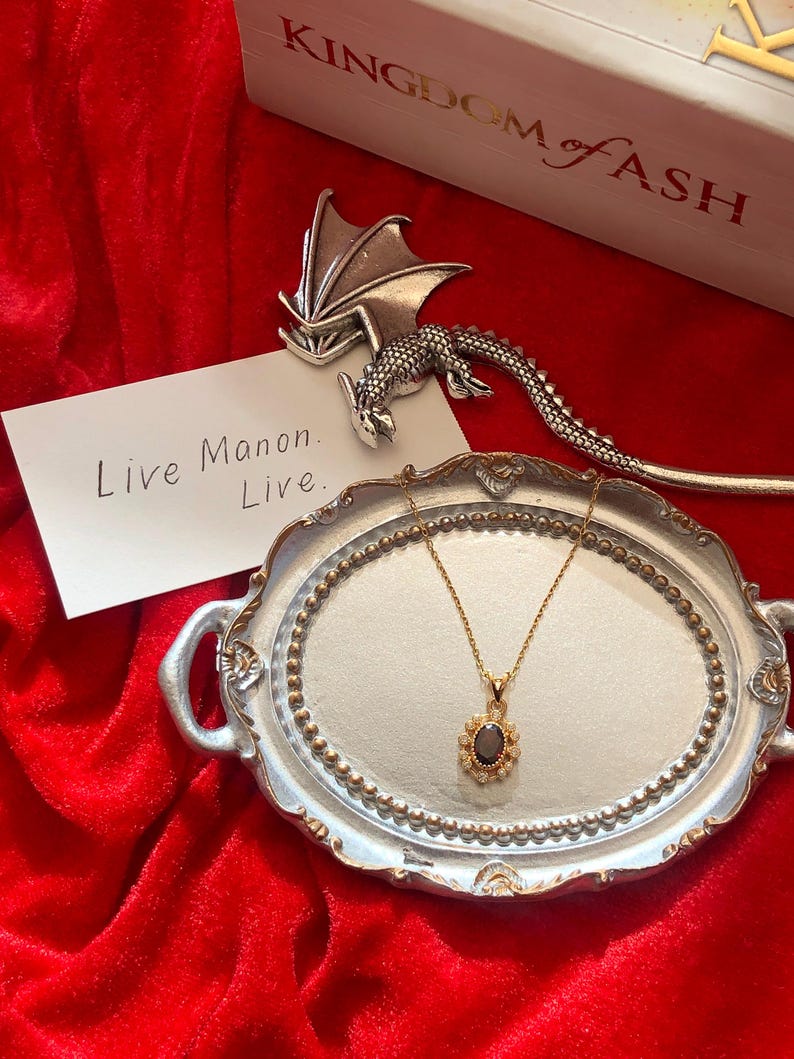 May include: A gold necklace with a dark oval pendant, surrounded by small white stones, rests on a silver tray. A silver dragon-shaped bookmark and a white card with the text "Live Manon. Live." are also present. The background is a red velvet surface.