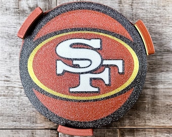 Puede incluir: Un posavasos redondo, rojo y negro, con el logotipo de los 49ers de San Francisco en el centro. El logotipo presenta las letras "SF" en blanco, con borde dorado, sobre un fondo rojo. El posavasos tiene tres pequeños mangos rojos.