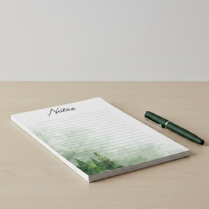May include: A notepad with the word "Notes" in cursive script, featuring a watercolor forest design at the bottom. A dark green pen rests on the right side of the notepad, which has lined pages.