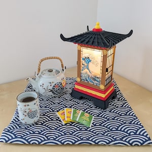Può includere: Una lampada in stile giapponese con tetto nero e base rossa, con un disegno a onde. Una teiera, una tazza da tè e bustine di tè sono su un tappetino a onde blu e bianco. La teiera e la tazza da tè hanno disegni di uccelli e fiori.