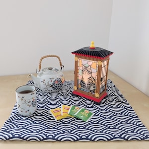 Puede incluir: Una linterna decorativa japonesa con un gato, efecto vidriera, una tetera, una taza de té y bolsitas de té sobre una esterilla con estampado de olas azules y blancas. La linterna tiene un techo negro y una base roja.