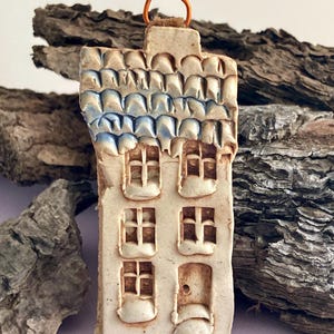 May include: A small ceramic house ornament with a blue and white tiled roof, white walls, and brown windows. The house has a small door and a loop at the top for hanging.