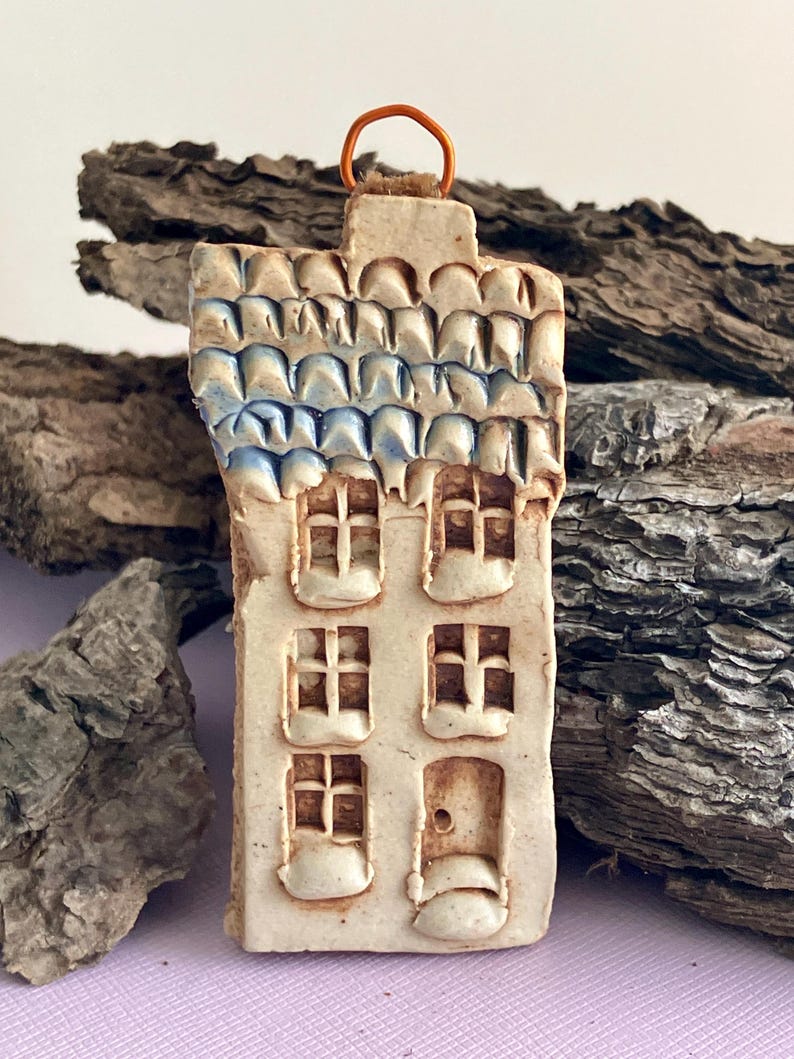 May include: A small ceramic house ornament with a blue and white tiled roof, white walls, and brown windows. The house has a small door and a loop at the top for hanging.