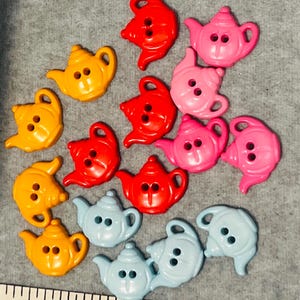 Könnte beinhalten: Eine Sammlung von teekannenförmigen Knöpfen in Rot, Orange, Pink und Hellblau, die jeweils zwei Löcher zum Annähen haben. Ein Teil eines weißen Maßbandes ist unten sichtbar und zeigt Zentimeter-Markierungen als Größenreferenz.