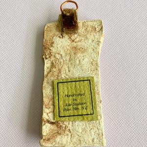 May include: Handcrafted by Julie Staddon, Palm Nth, N.Z.  A small, rectangular, brown and white textured wall hanging with a copper wire loop and a brown velvet hanger.