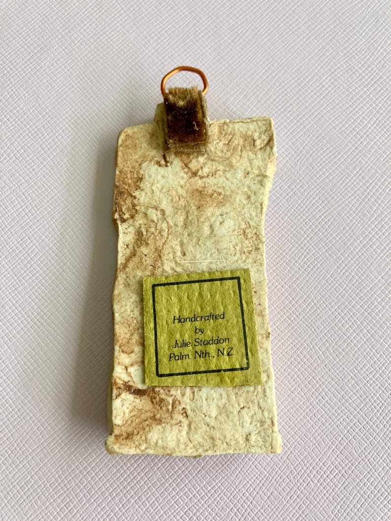 May include: Handcrafted by Julie Staddon, Palm Nth, N.Z.  A small, rectangular, brown and white textured wall hanging with a copper wire loop and a brown velvet hanger.