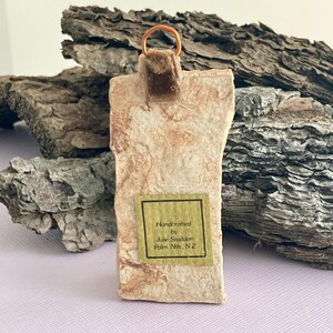 May include: Handcrafted ceramic ornament with a brown velvet ribbon and a gold wire hanger. The ornament has a light brown and white marbled appearance. A small label on the ornament reads "Handcrafted by Julie Staddon Palm Nth, N.Z."