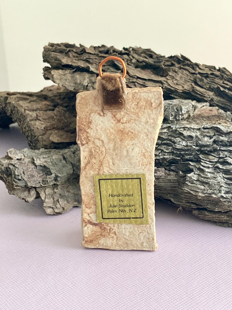 May include: Handcrafted ceramic ornament with a brown velvet ribbon and a gold wire hanger. The ornament has a light brown and white marbled appearance. A small label on the ornament reads "Handcrafted by Julie Staddon Palm Nth, N.Z."