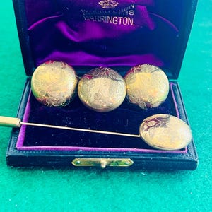 May include: A set of three round gold-toned buttons and a matching stick pin, each featuring an intricate floral engraving. The items are presented inside a black hinged box with a purple velvet interior lining and the text W.H.H. & SONS WARRINGTON.