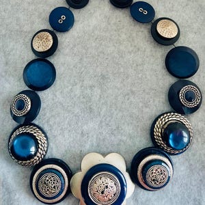 Könnte beinhalten: Eine auffällige Halskette aus verschiedenen marineblauen und silbernen Knöpfen. Das Design umfasst eine Mischung aus glatten, strukturierten und metallischen Knöpfen, die kreisförmig angeordnet sind. Ein großer, blütenförmiger Knopf bildet den Mittelpunkt.