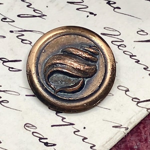 May include: A circular, antique copper button featuring a raised seashell design. The button has a dark, aged patina and is set against a backdrop of handwritten script on aged paper.