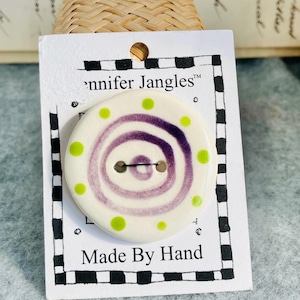 May include: A handmade, cream-coloured button featuring a purple spiral and green polka dots. The button is on a card with the text "Jennifer Jangles" and "Made By Hand" within a black and white chequered border.