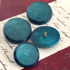 Könnte beinhalten: Vier runde, teal-farbene Knöpfe mit glänzendem Finish. Ein Knopf hat einen goldfarbenen Schaft. Die Knöpfe sind auf einem Stück Vintage-Papier mit handschriftlichem Schriftzug angeordnet. Die Knöpfe sind tief teal.