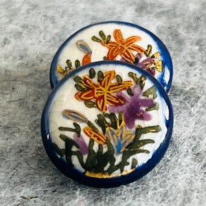 Könnte beinhalten: Zwei runde Keramikknöpfe mit weißem Craquelé-Glasur-Hintergrund und handgemalten Blumenmotiven in Orange, Violett und Grün. Jeder Knopf hat einen tiefblauen Rand und eine glänzende Oberfläche, die detaillierte botanische Illustrationen zeigt.