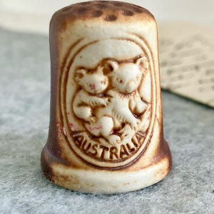 May include: A vintage thimble with a cream-coloured design of two koalas embracing. The thimble has a brown rim and the word "AUSTRALIA" is written around the koalas. A collectible item.