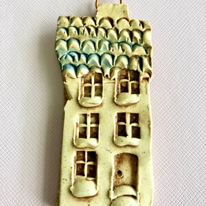 May include: A ceramic house ornament with a blue and white roof, three windows, and a door. The ornament is white with brown accents and has a loop for hanging.