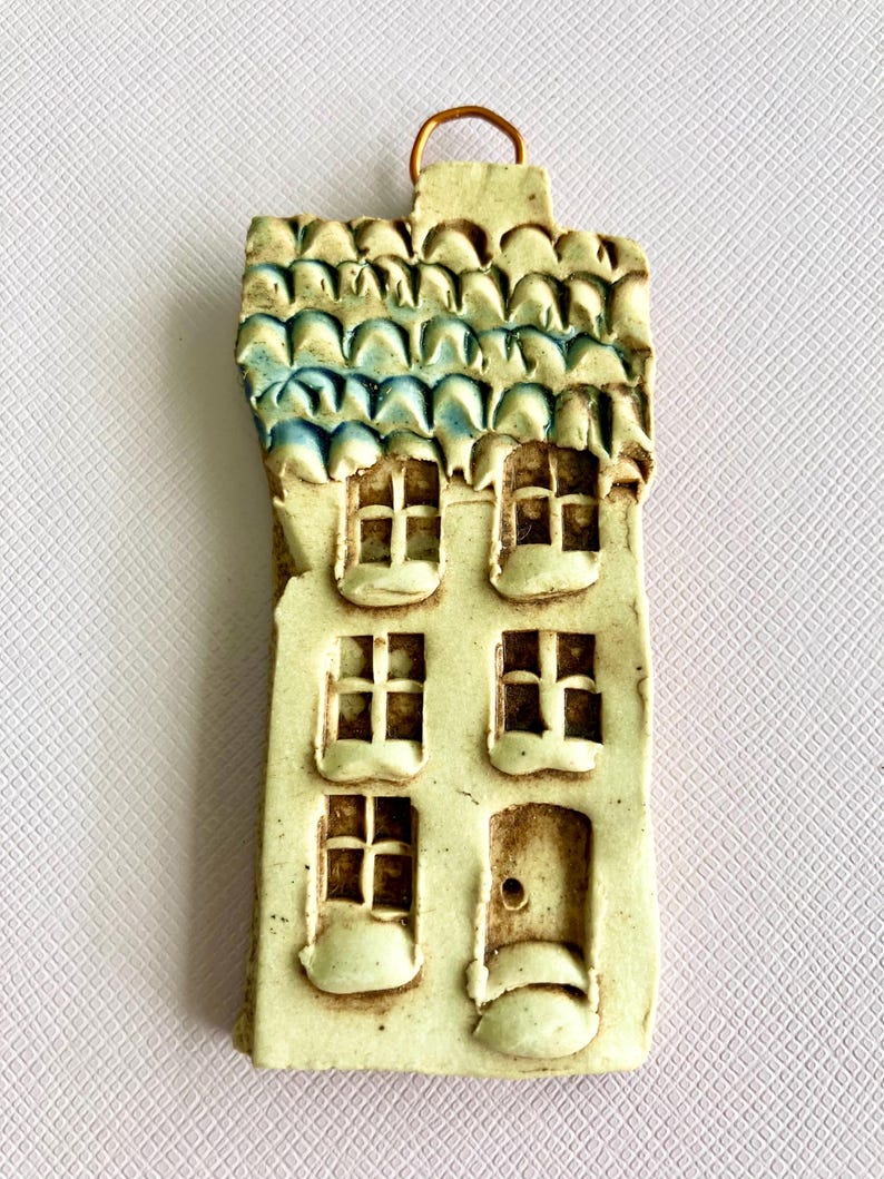 May include: A ceramic house ornament with a blue and white roof, three windows, and a door. The ornament is white with brown accents and has a loop for hanging.