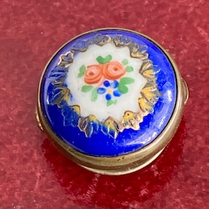 May include: A small, round, blue and white enamel box with a floral design. The box has a gold trim and is decorated with a white enamel centre featuring a hand-painted floral design of pink roses and blue flowers.