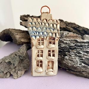 May include: A small ceramic house ornament with a blue and white tiled roof, white walls, and a small door. The ornament has a copper wire loop for hanging.